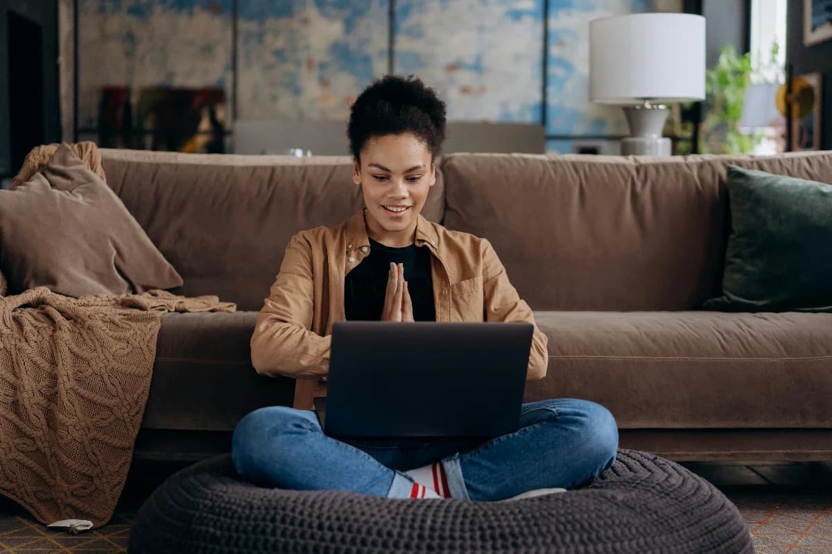 Woman sitting cross-legged at home, smiling while using a laptop and making a grateful hand gesture